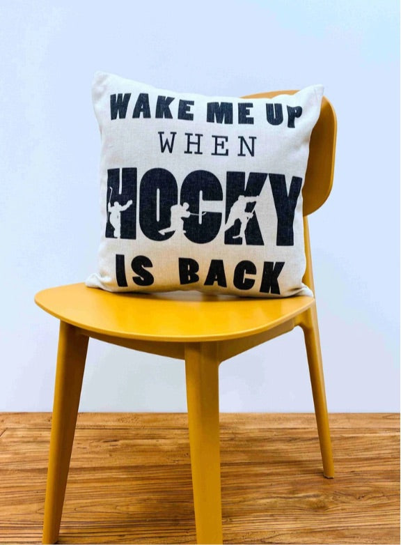 A close-up image of a beige throw pillow with the words "Hockey Is Back" displayed in bold typography. The pillow features a canvas-touch fabric and a hidden zipper closure for easy cleaning. It is an ideal decorative accent for hockey enthusiasts, offering both style and comfort. Dimensions are 17x17"